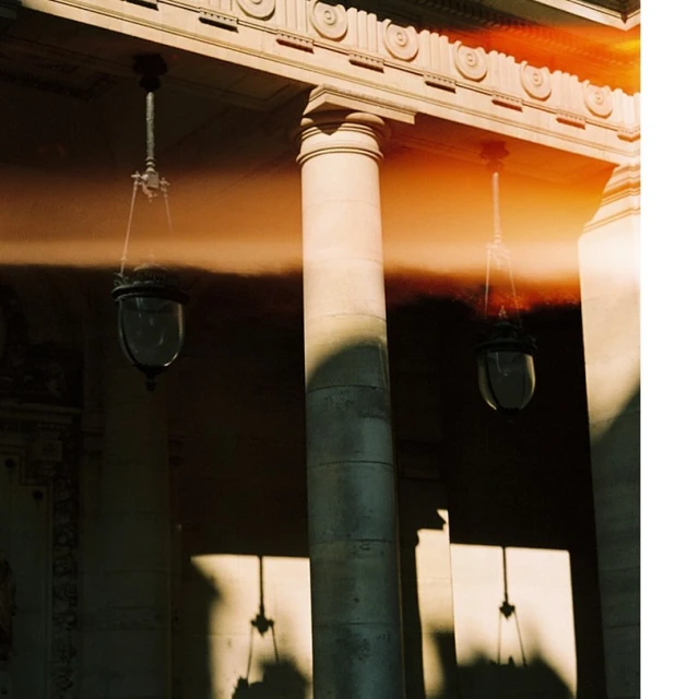 Exterior of a building with classical architecture featuring columns and hanging lamps in soft, natural light casting shadows.