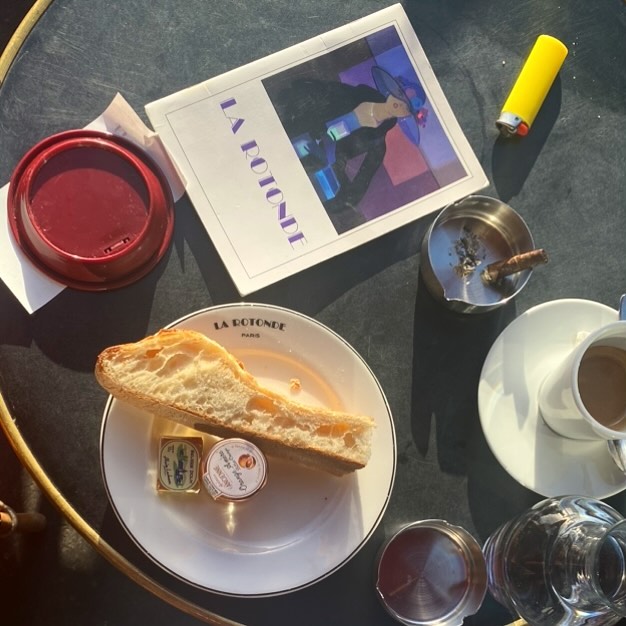 Overhead shot of a table featuring a plate with a bread slice and condiments, a coffee, a magazine, and an ashtray with a cigarette.