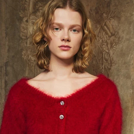 Close-up portrait of a young woman with wavy blonde hair wearing a red textured cardigan against a muted patterned wall.