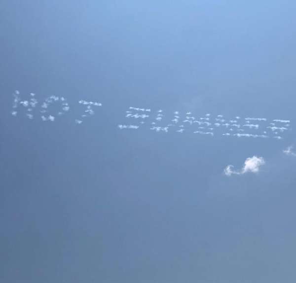 Skywriting in a blue sky that spells out 'HOT SUMMER' with puffy white clouds. The sky is a clear, vibrant blue, with only a few small clouds.