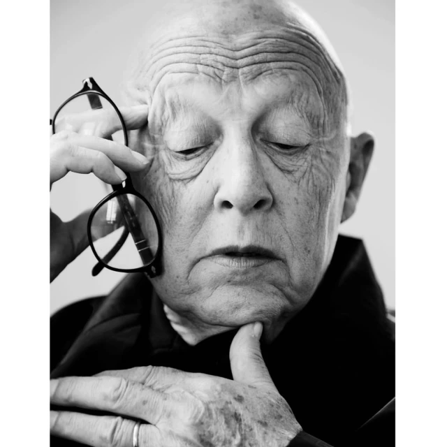 Black and white portrait of an elderly adult male with eyeglasses, eyes closed, resting his chin on his finger against a plain background.