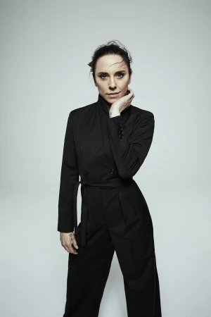 Portrait of a woman wearing a dark belted jumpsuit with her hand near her chin against a light background.