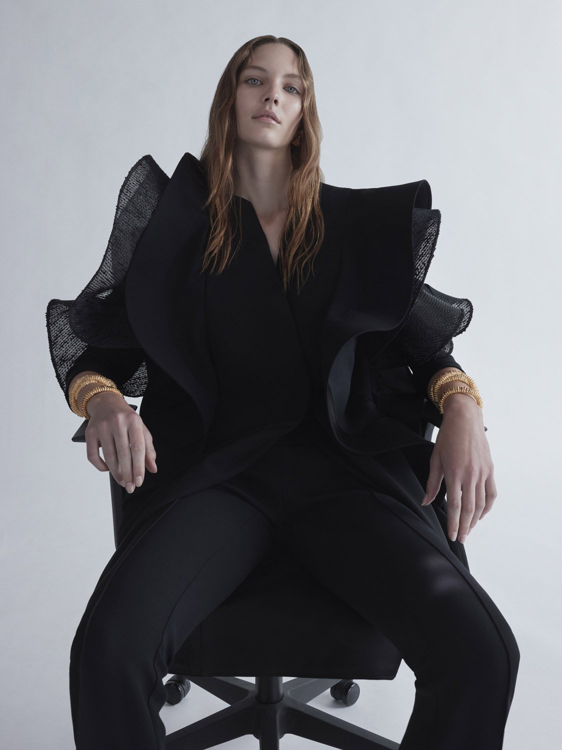 Fashion portrait of a model wearing a black ruffled jacket and pants, seated on an office chair against a plain background.