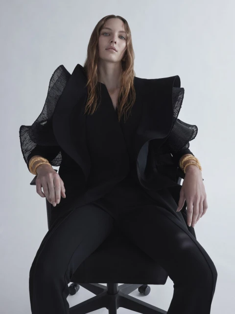 Fashion portrait of a model wearing a black ruffled jacket and pants, seated on an office chair against a plain background.
