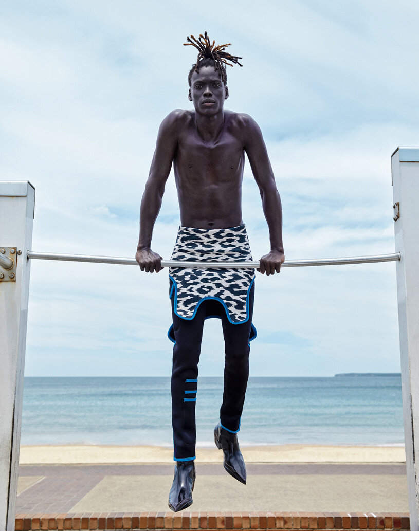 Athletic Black man with dreadlocks works out on a bar on a beach, wearing fashionable designer clothes with the ocean in the background.