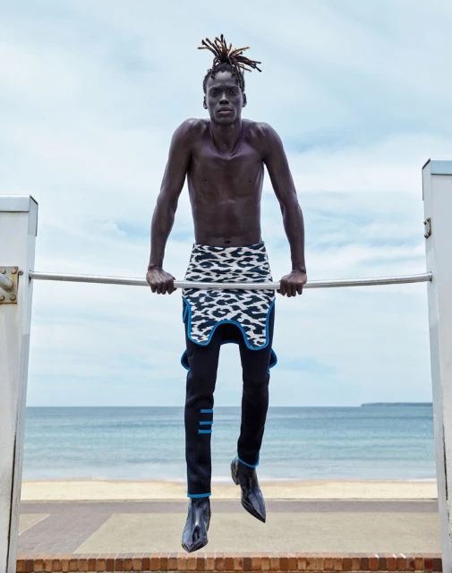 Athletic Black man with dreadlocks works out on a bar on a beach, wearing fashionable designer clothes with the ocean in the background.