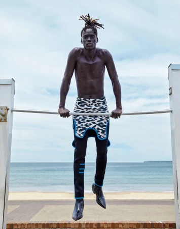 Athletic Black man with dreadlocks works out on a bar on a beach, wearing fashionable designer clothes with the ocean in the background.