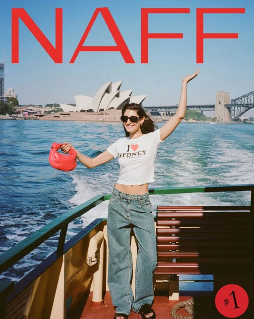 Woman poses on ferry with Sydney Opera House backdrop, wearing casual tee, jeans, holding a red purse, enjoying the sunny day.