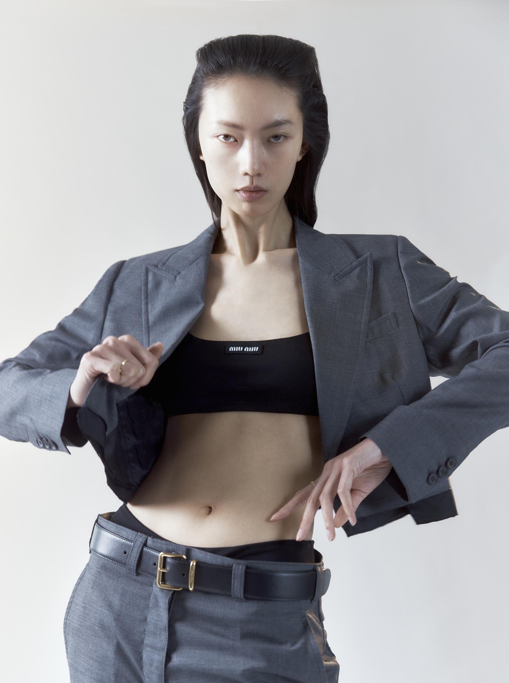 Asian fashion model in a grey blazer, black top, and grey pants poses indoors against a neutral background in a studio setting.