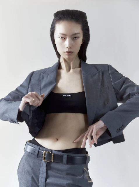 Asian fashion model in a grey blazer, black top, and grey pants poses indoors against a neutral background in a studio setting.