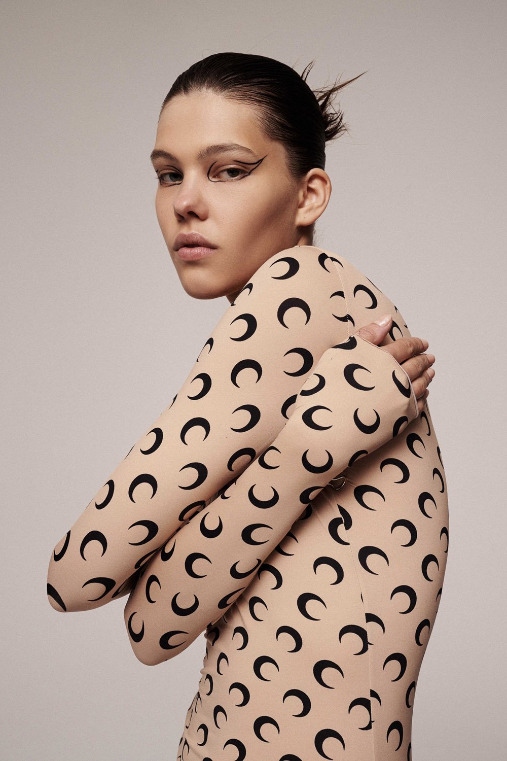 Close-up of a female model in a nude bodysuit with black crescent moon patterns, striking a pose against a neutral backdrop.
