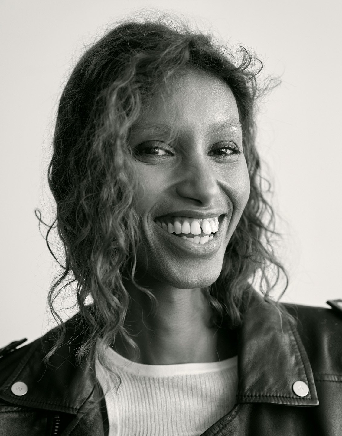 Portrait of a smiling woman with curly hair wearing a black leather jacket and white top against a white background