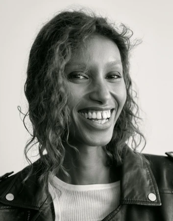 Portrait of a smiling woman with curly hair wearing a black leather jacket and white top against a white background