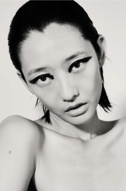 Striking black and white portrait of an Asian woman with slicked-back hair and bold, dark winged eyeliner, against a pale background.