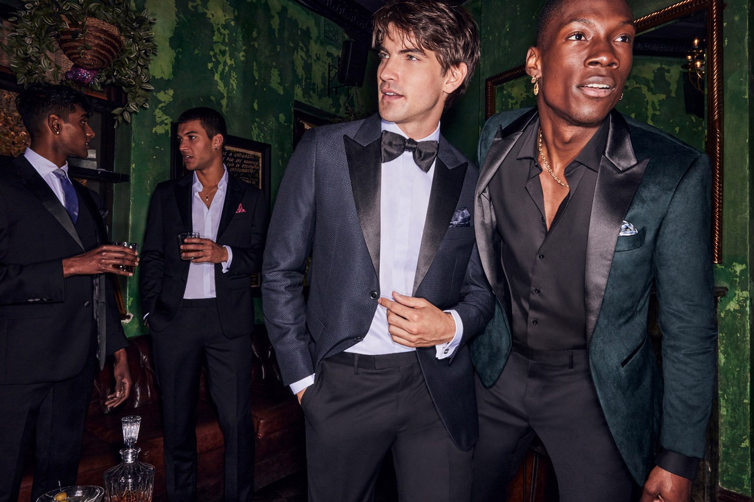 Group of four men in formal wear gather indoors at a social event, wearing suits and tuxedos, drinking, and talking in a bar setting.