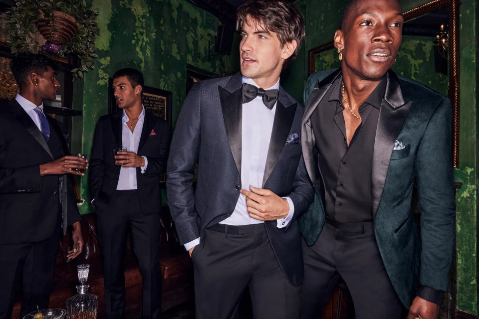 Group of four men in formal wear gather indoors at a social event, wearing suits and tuxedos, drinking, and talking in a bar setting.