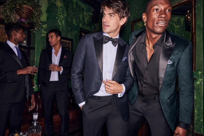 Group of four men in formal wear gather indoors at a social event, wearing suits and tuxedos, drinking, and talking in a bar setting.