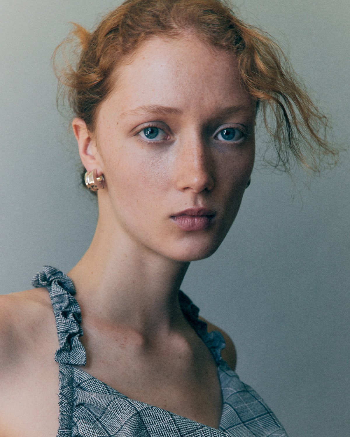Close-up portrait of a fair-skinned young woman with red hair and blue eyes, wearing a gray plaid top against a neutral background.