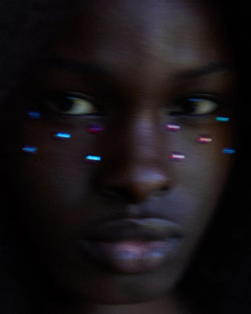 Close-up portrait of an adult woman with dark skin, adorned with blue and pink dots around her eyes in soft, low-key lighting.