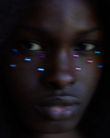 Close-up portrait of an adult woman with dark skin, adorned with blue and pink dots around her eyes in soft, low-key lighting.