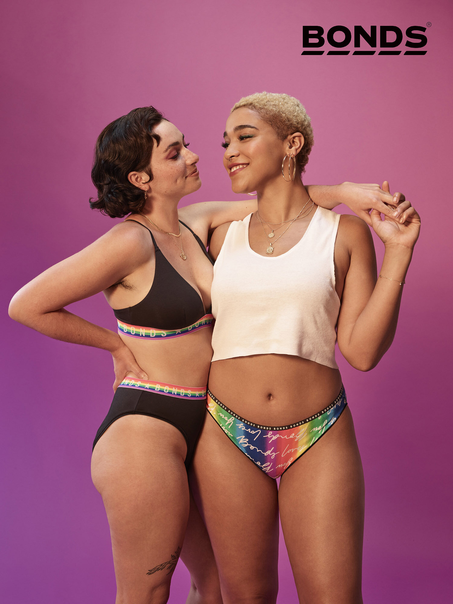 Two smiling women pose together indoors in Bonds underwear; one wears a bralette and briefs set, the other a cropped tank top and briefs.