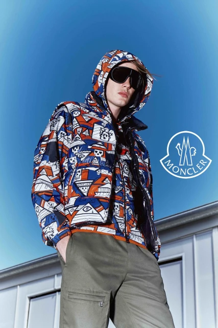 Fashion shot of a man wearing a patterned Moncler hooded jacket, sunglasses, and olive green pants against a bright blue sky backdrop.