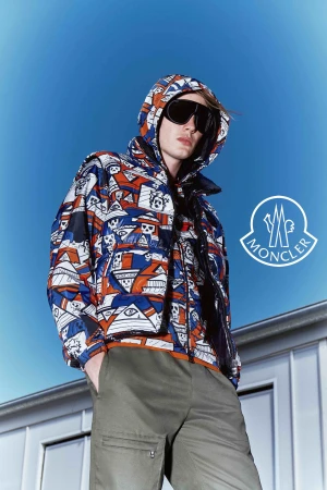 Fashion shot of a man wearing a patterned Moncler hooded jacket, sunglasses, and olive green pants against a bright blue sky backdrop.