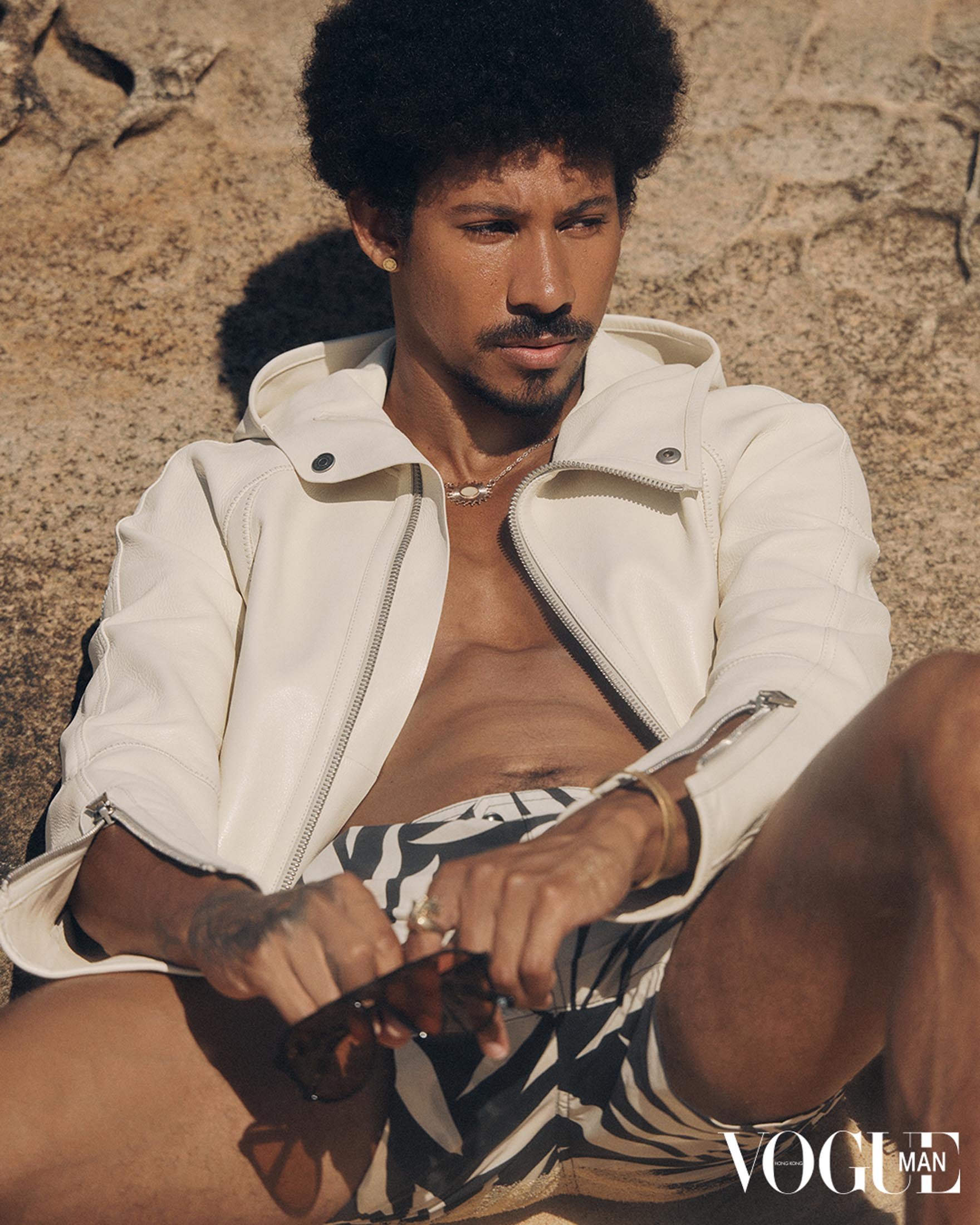 A stylish man with an afro, wearing a white jacket and patterned shorts, sits on a sandy beach near a rocky outcrop.