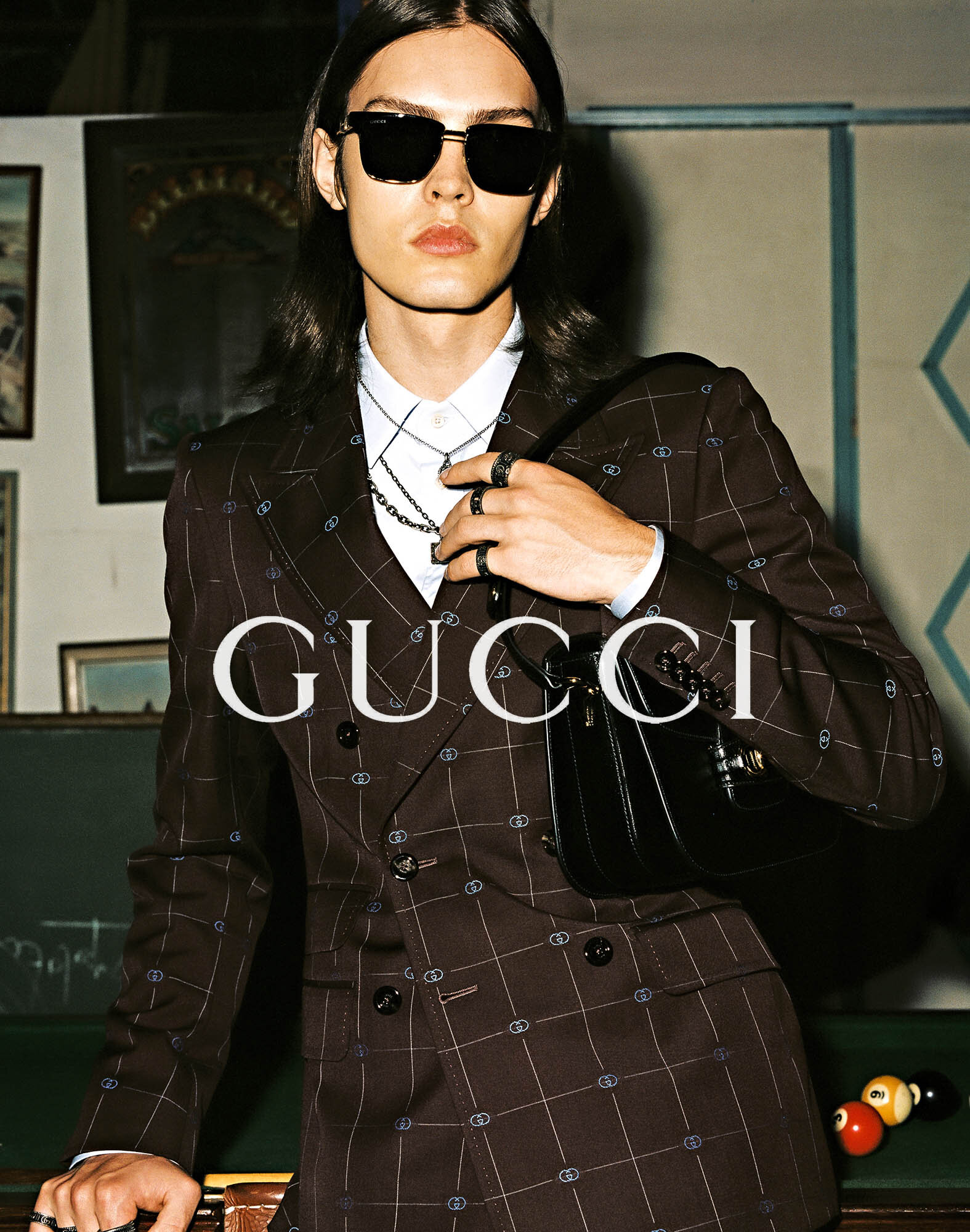 An adult male poses wearing a brown double-breasted suit, jewelry, and black sunglasses, leaning on a pool table with Gucci branding.