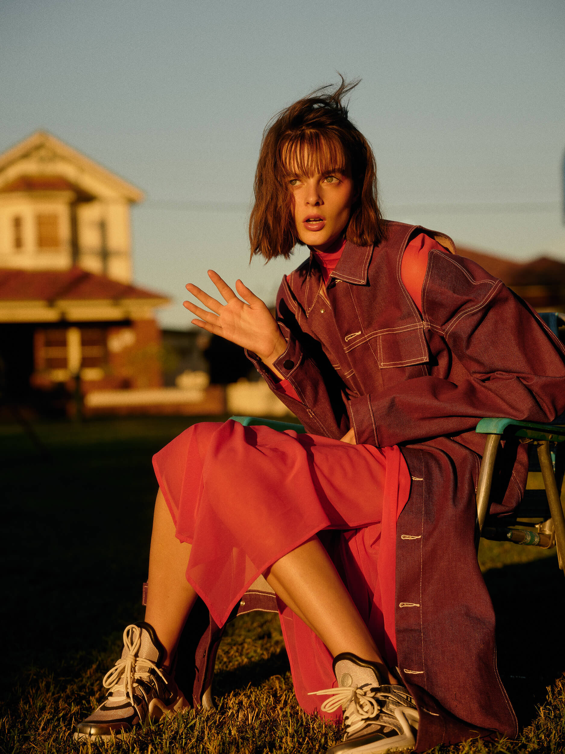Female fashion model with short brown hair poses on a lawn chair wearing a jacket and skirt in a field, house in the background.