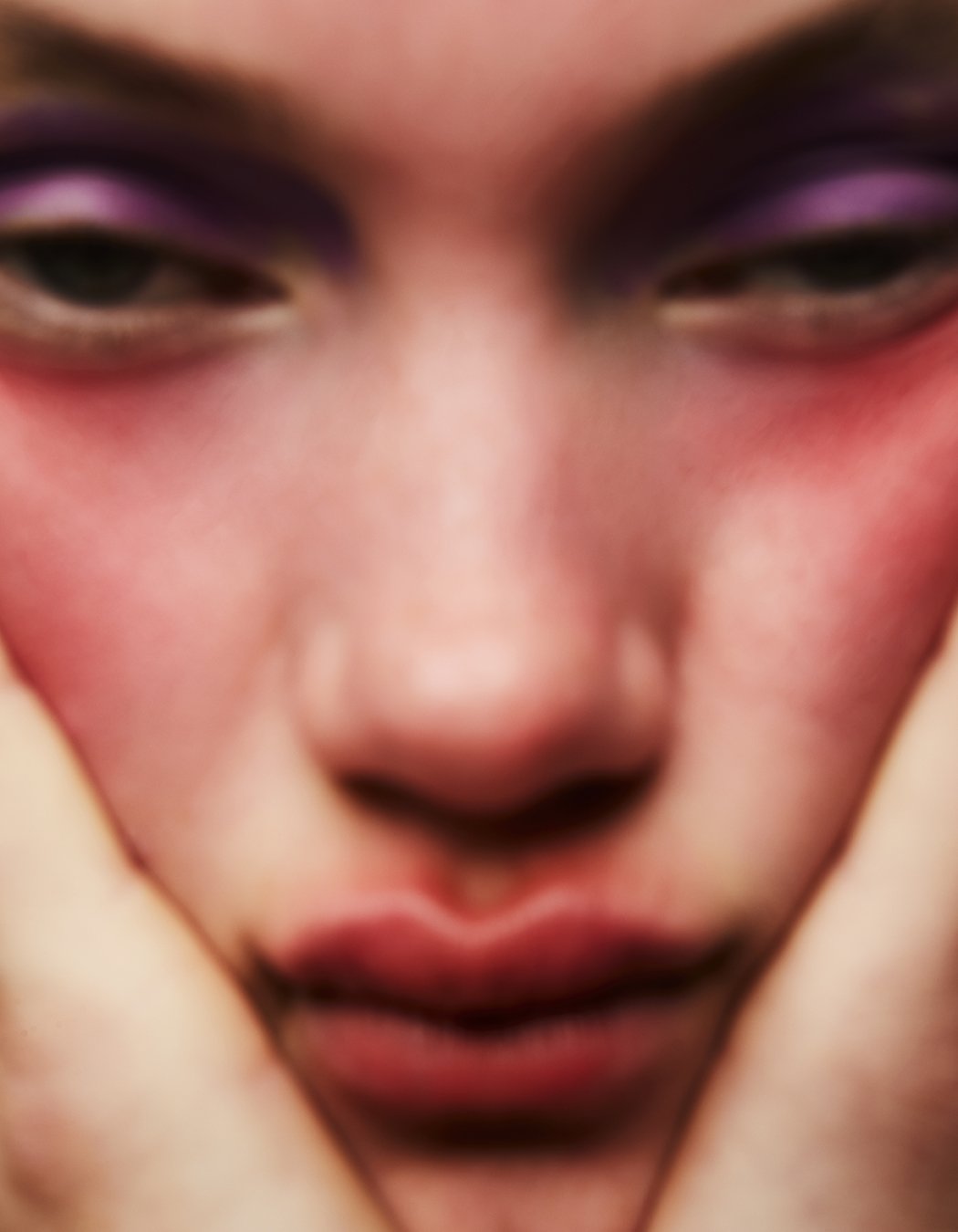 Close-up portrait of a fair-skinned person with purple eyeshadow and reddish blush holding their face with hands, soft focus.