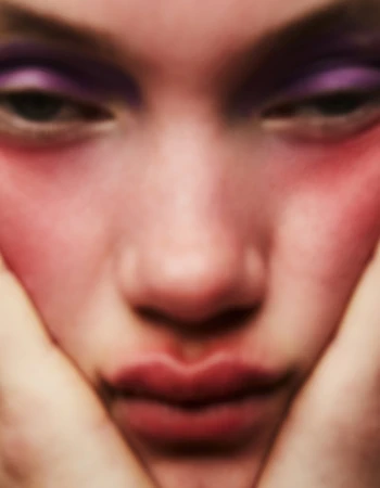Close-up portrait of a fair-skinned person with purple eyeshadow and reddish blush holding their face with hands, soft focus.