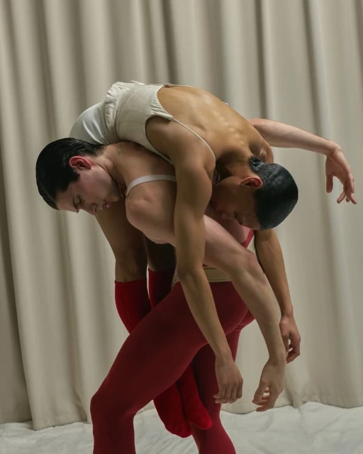 Two athletic male ballet dancers in red leggings with white leotards, one carrying the other on his shoulders against a cloth backdrop.
