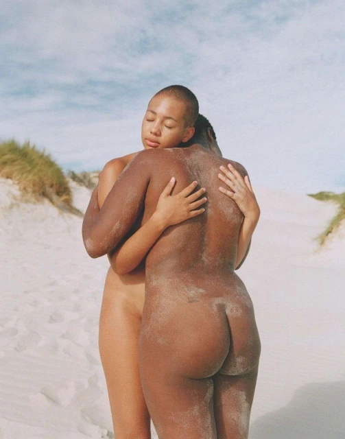 Two nude adult figures embrace outdoors on a sandy dune under a bright sky; one person faces the camera in a close-up shot.