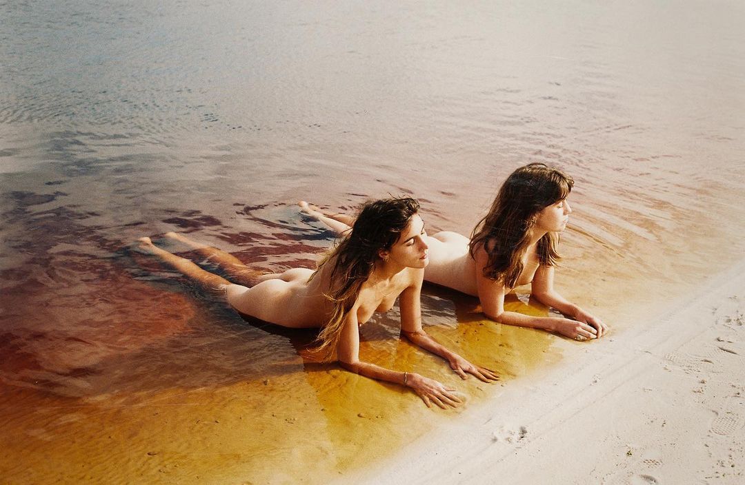 Two women lie side-by-side on a sandy beach at the edge of shallow water, basking in sunlight. The water is tinted with reddish hues.