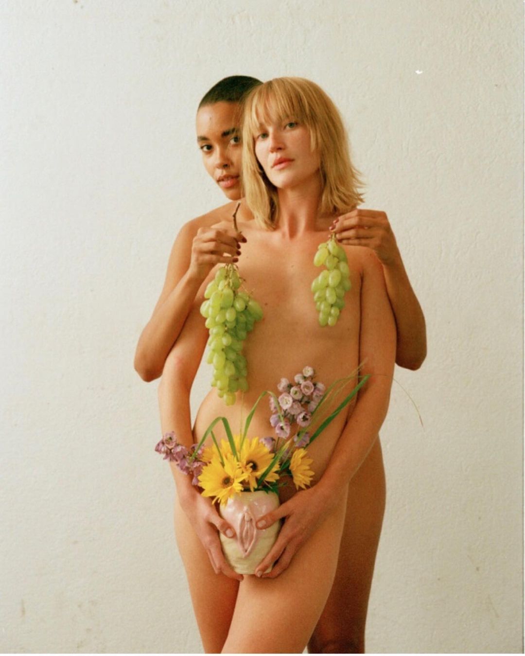 Two figures pose creatively, one with short blonde hair and one with a shaved head, decorated with grapes and flowers against a white background.