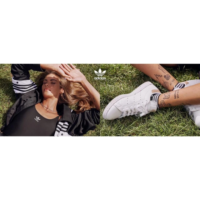 Two women relax on grass wearing Adidas apparel and Stan Smith shoes. The Adidas logo is visible in the center of the frame.