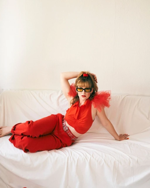 Stylish woman in red outfit and yellow sunglasses lounges on white couch in studio, hair styled with red heart clip, relaxed pose.