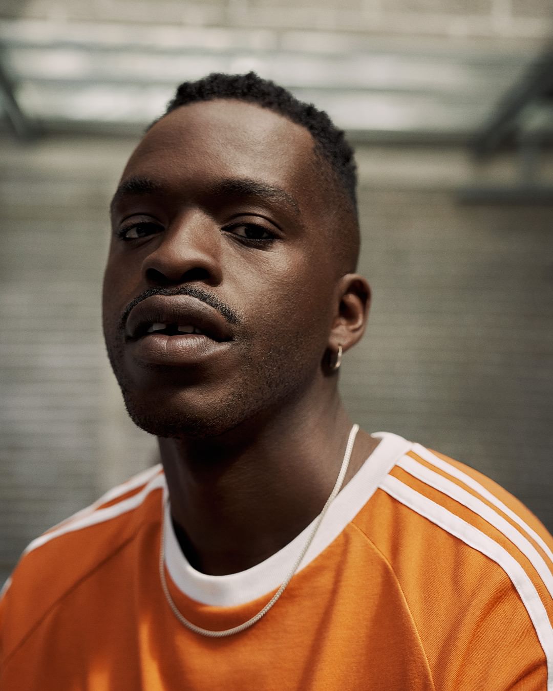 Close-up of a young Black man with short hair and a mustache, wearing a bright orange t-shirt with white stripes and a silver chain necklace.