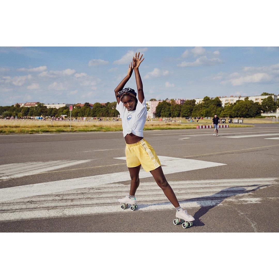 A young, dark-skinned woman roller skating on an airstrip, wearing yellow shorts and a white t-shirt, with a blue sky and clouds.
