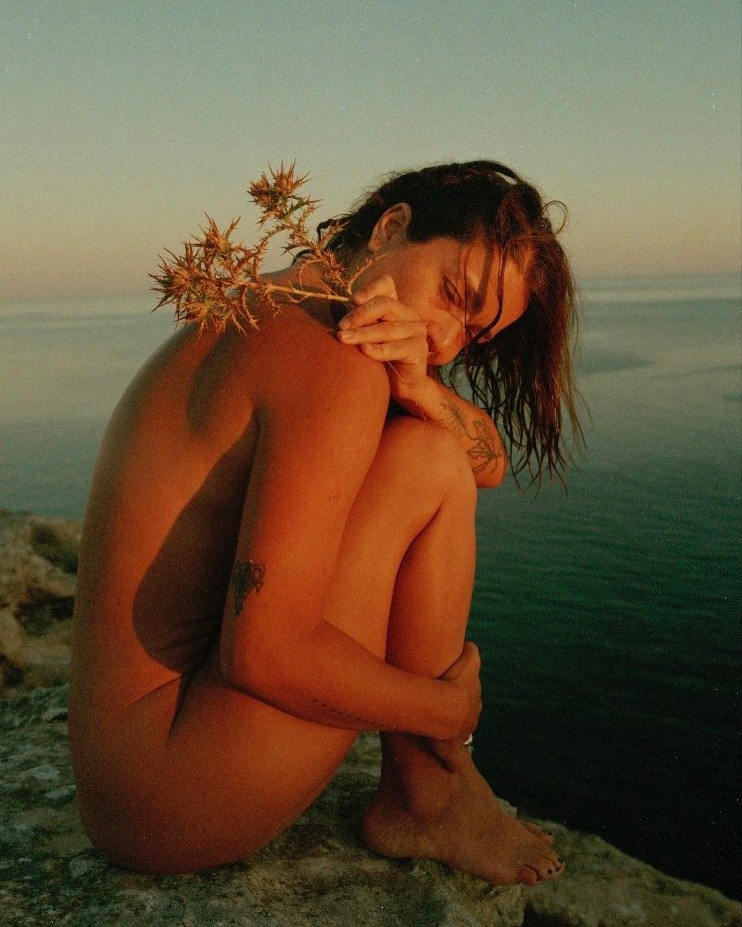 Woman with bare skin sits on a rock near the ocean, holding a thistle; warm sunset light cast on skin and sea in the background.
