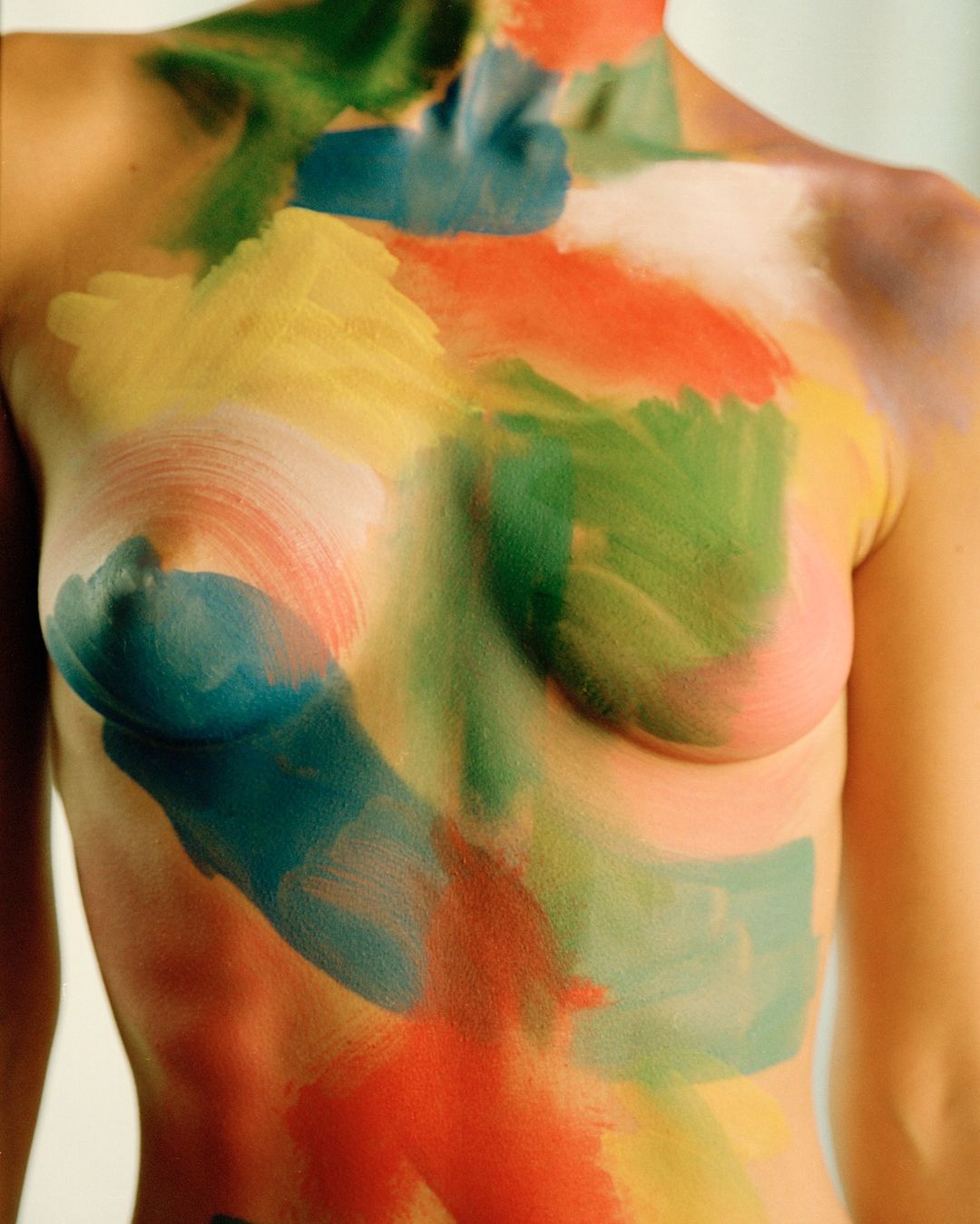 Conceptual image of a torso covered in swathes of blue, green, yellow, and red paint against a white background in a medium shot.
