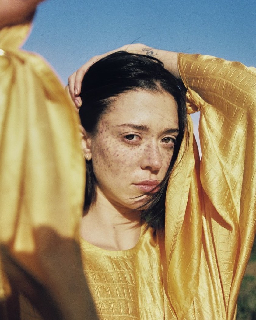 Close-up portrait of a young adult female with dark hair and freckles, wearing a yellow textured dress, against a blue sky backdrop.