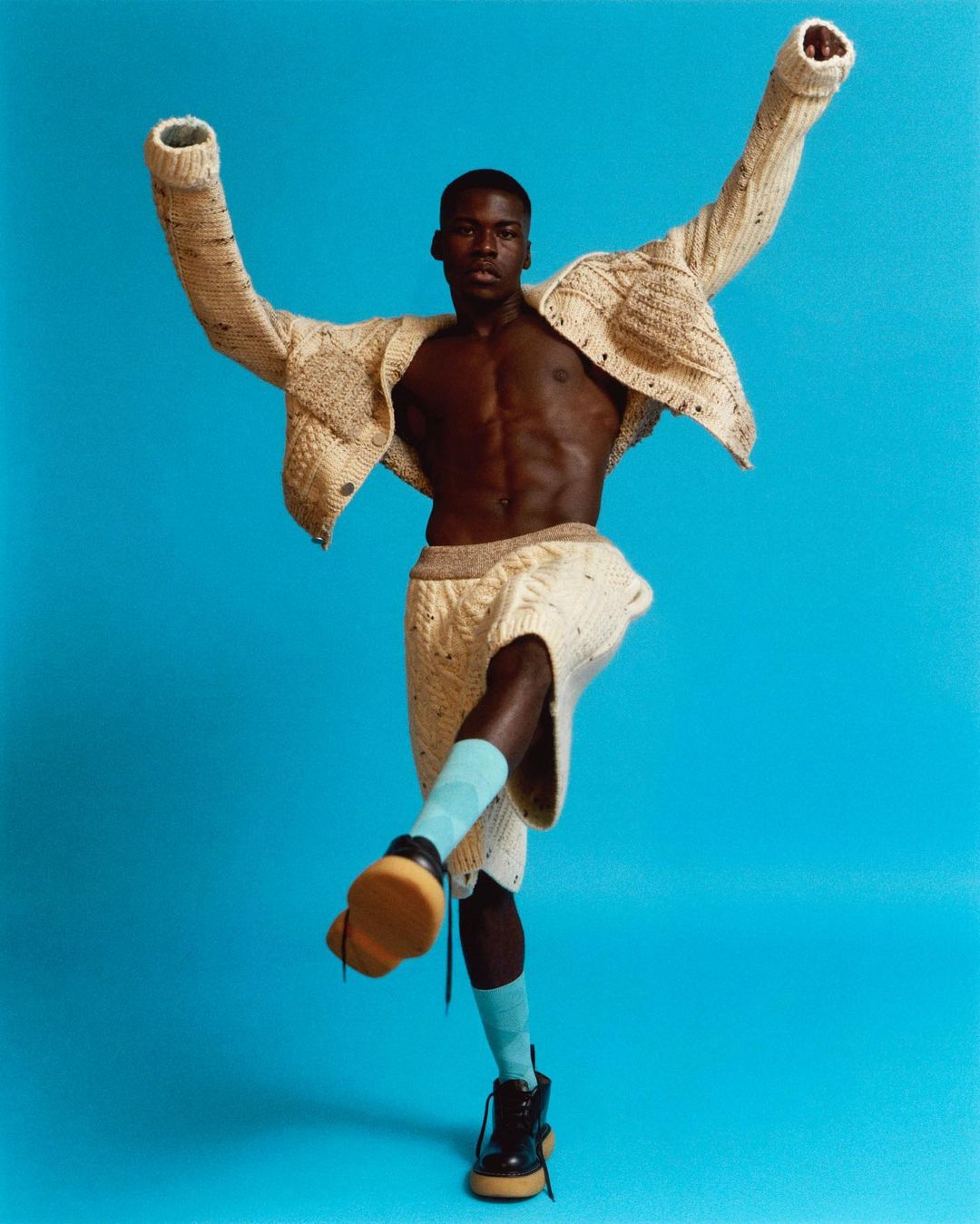 Athletic build adult male strikes a dynamic pose with a turquoise backdrop, wearing a knitwear set, blue socks, and chunky sole boots in a studio setting.