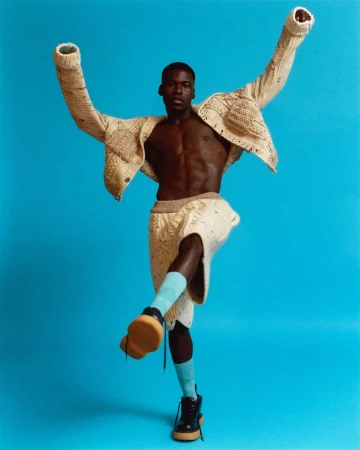 Athletic build adult male strikes a dynamic pose with a turquoise backdrop, wearing a knitwear set, blue socks, and chunky sole boots in a studio setting.