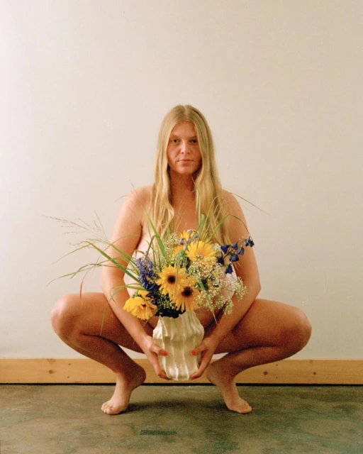 An adult woman squats indoors holding a bouquet of sunflowers and purple flowers in an off-white vase against a neutral wall.