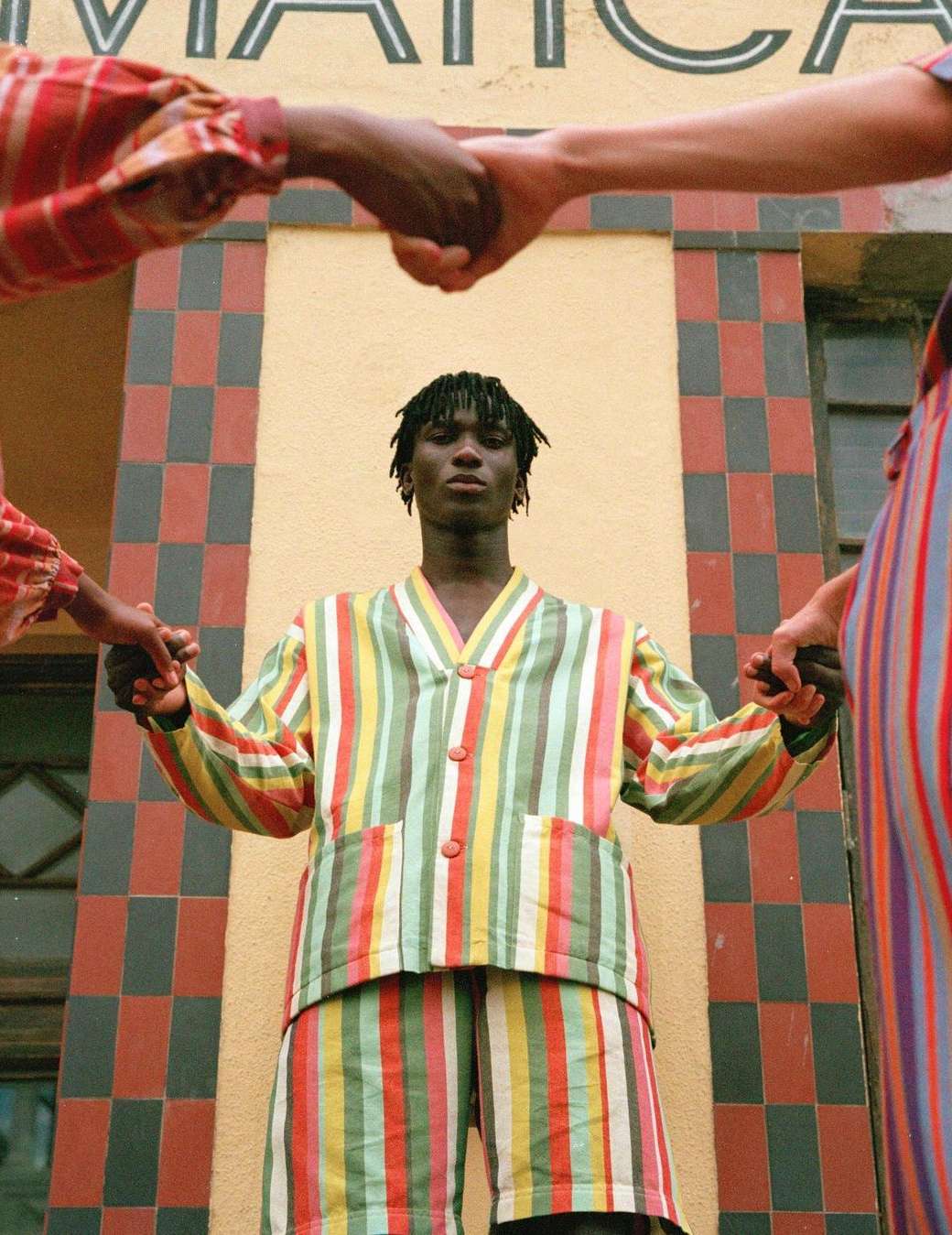 A person in a striped outfit stands in front of a building with tiled walls while two other people hold their hands, creating a circle.