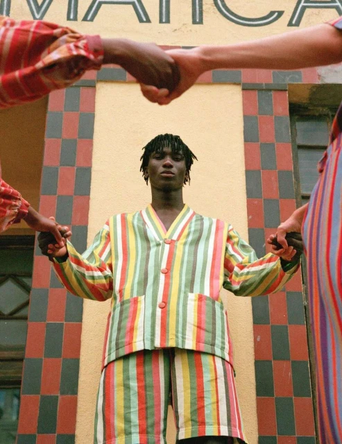 A person in a striped outfit stands in front of a building with tiled walls while two other people hold their hands, creating a circle.