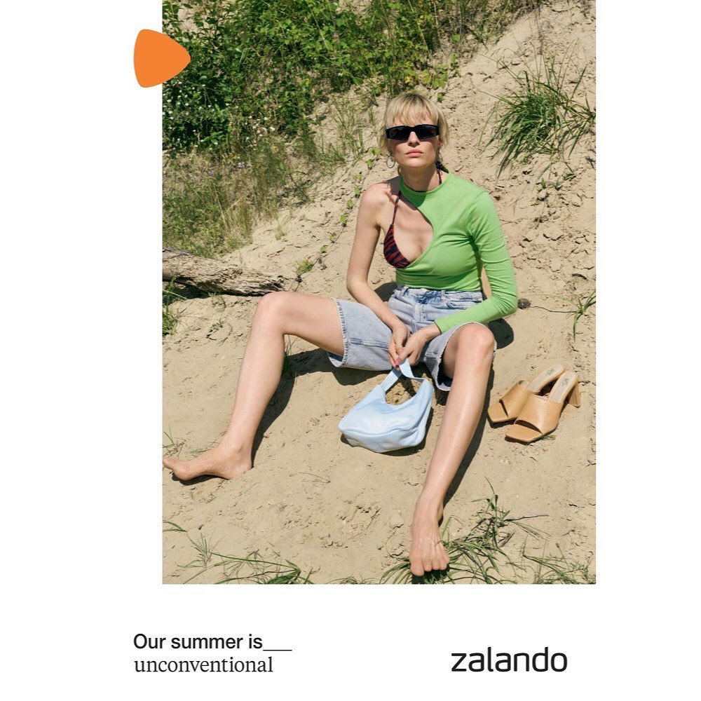A woman sits in sand wearing a green, long-sleeved top, jean shorts, and sunglasses, holding a light-blue bag with tan heels nearby.