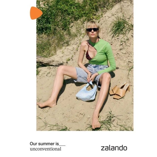 A woman sits in sand wearing a green, long-sleeved top, jean shorts, and sunglasses, holding a light-blue bag with tan heels nearby.